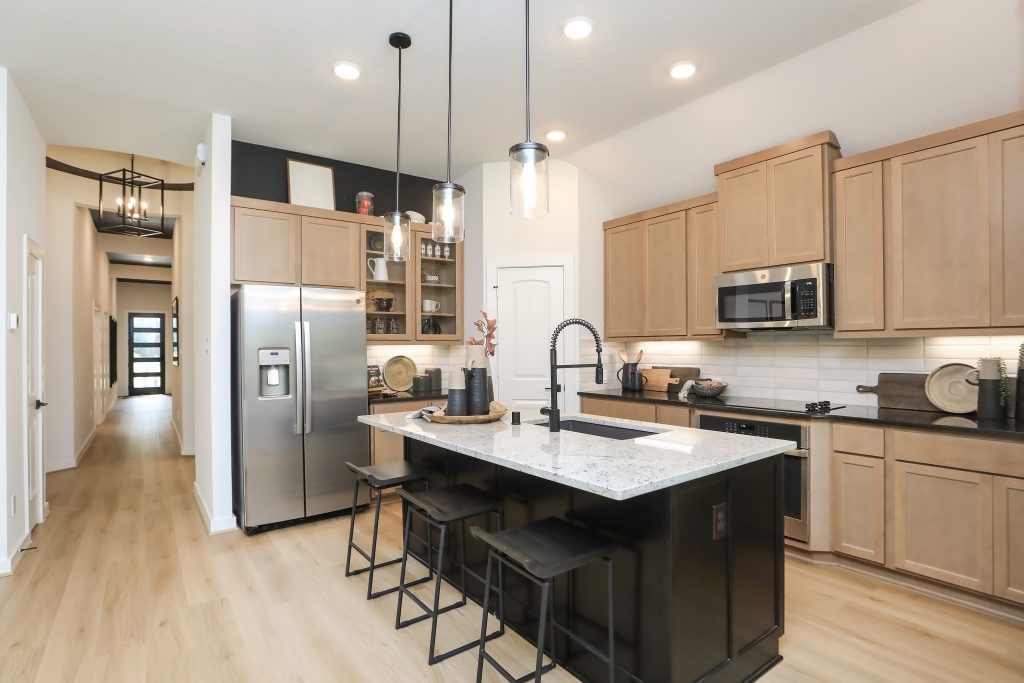 Modern kitchen with light wood cabinets, stainless steel appliances, a large island with a sink and black stools, pendant lights above, and light wood flooring; hallway visible in the background.
