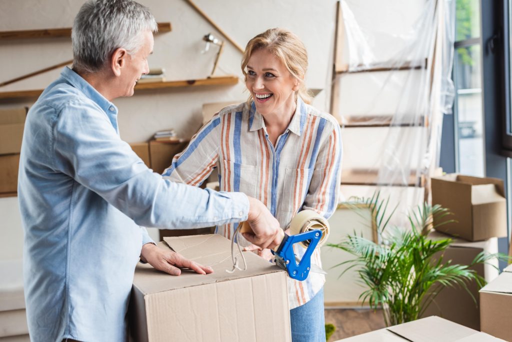 A smiling man and woman pack cardboard boxes together, sealing one with packing tape in a bright room filled with moving boxes and houseplants.