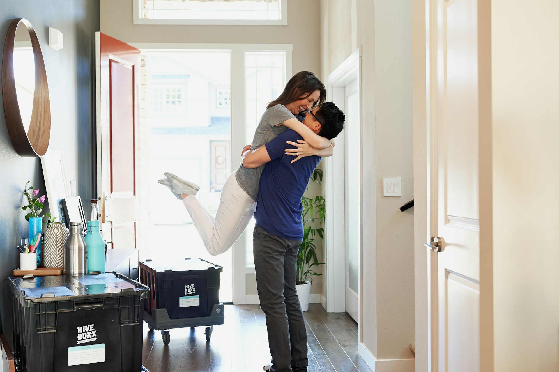 A man lifts and hugs a woman in a bright hallway beside black moving boxes, suggesting they are moving into a new home. Both appear happy, with sunlight streaming through the windows.